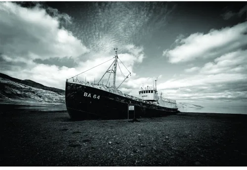 A black and white photograph of an abandoned fishing trawler named DA 64 beached on dark sand under a dramatic cloudy sky.
