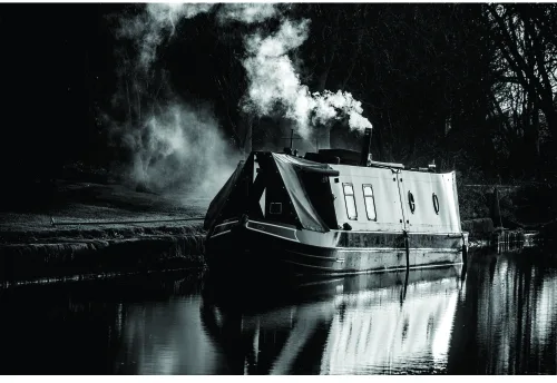 A black-and-white photograph of a narrowboat on a canal with large plumes of steam rising from its chimney, reflected perfectly in the still water below.