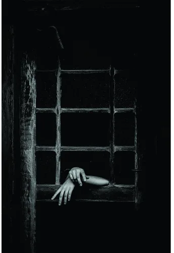 Black and white photograph of two hands reaching through a weathered barred window set into a rough stone wall.