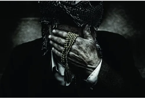 Black and white photograph of aged hands held together in prayer position, wearing rings and holding a beaded strand.