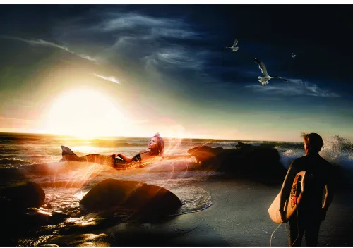 A surfer in a wetsuit stands on a rocky beach holding a surfboard, gazing at a dramatic golden sunset with waves and boulders in the foreground.