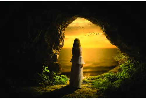 A woman in dark clothing stands silhouetted inside a cave, facing a bright golden sunset visible through the cave opening, with green moss covering the rocky interior.