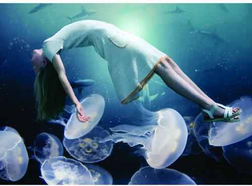 A woman in light-coloured clothing floats upside down underwater, surrounded by glowing jellyfish with translucent bells and trailing tentacles in shades of blue and white.