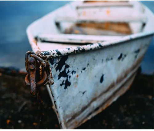 Photograph of a weathered white fishing boat with rust-stained rope and hardware, with a second boat blurred in the background against blue water.