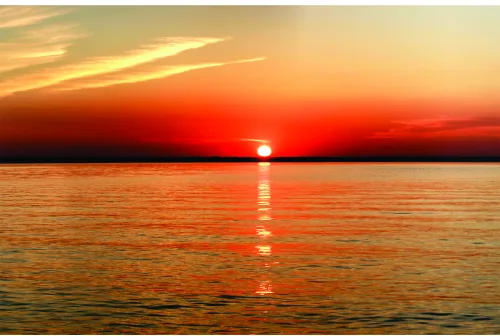 A photographic image of a large sun on the horizon reflecting onto calm water at sunset, with golden and orange tones dominating the sky.