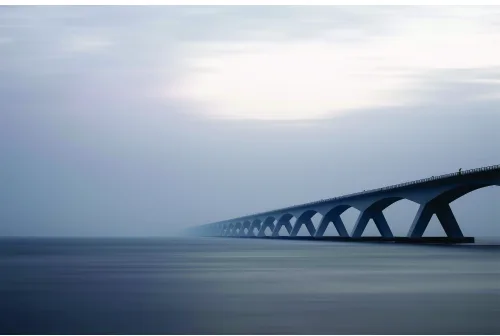 A long concrete bridge with repeating arch supports extends across still blue-grey water under an overcast sky.