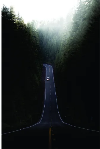 Aerial view of a narrow asphalt road stretching straight through dense green forest, with a single dark vehicle in the distance and filtered sunlight breaking through the canopy.