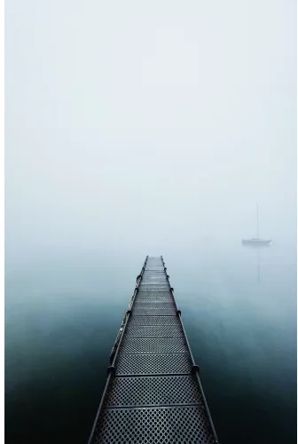 A long metal pier with geometric mesh flooring extends straight into dense fog over calm water, with a sailboat barely visible in the mist.