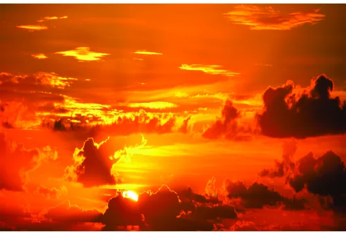 A dramatic sunset photograph showing dark silhouetted clouds against a fiery sky of orange, yellow and red light.