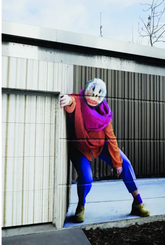 A street art mural of a skateboarder wearing a purple helmet and colourful athletic wear in a low crouch, hand extended, against an urban industrial backdrop.