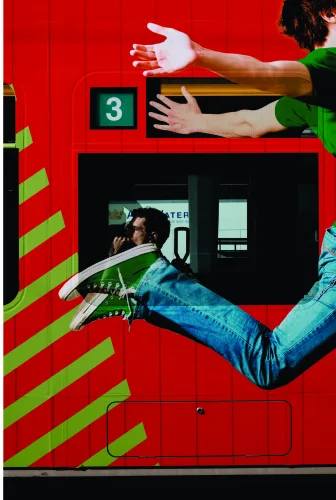 Collage artwork of a person in casual clothes climbing a bright red wall marked with green diagonal stripes, surrounded by fragmented urban imagery.