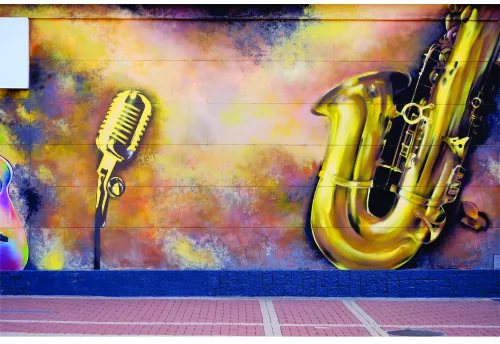 Street art mural showing a golden vintage microphone on the left and a golden saxophone on the right against a textured background of warm yellows, oranges and cool purples and blues.