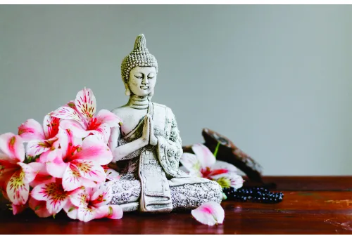 A white Buddha statue sits among bright pink flowers and dark incense sticks on a wooden surface against a grey background.