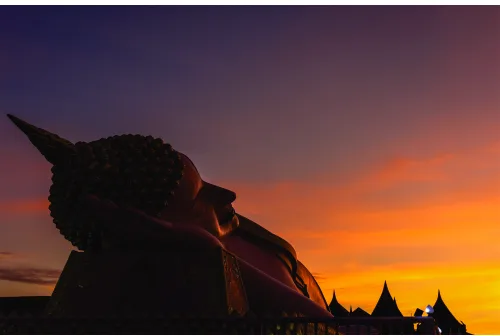 A dark Buddha statue in profile is backlit by a golden orange and amber sunset sky with deep blue shadows.