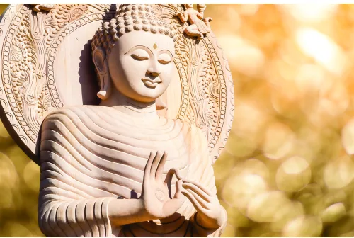 A cream-coloured Buddha statue with closed eyes and serene expression, wearing robes with linear carving details, framed by an ornate circular halo, photographed in warm golden sunlight.