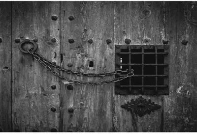 Black and white photograph of a weathered wooden door with scattered nail holes, a chain strung diagonally across it, and a small barred window.