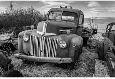 Black and white photograph of an abandoned vintage truck partially overgrown with beach grass, with driftwood and other discarded vehicles visible in the background.