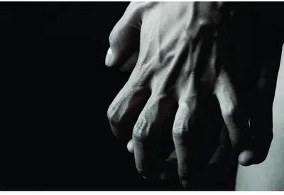 Photographic image of two pale hands reaching downward against a black background, rendered in monochrome with dramatic shadows.