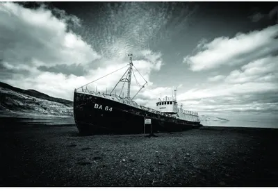 A black and white photograph of an abandoned fishing trawler named DA 64 beached on dark sand under a dramatic cloudy sky.