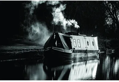 A black-and-white photograph of a narrowboat on a canal with large plumes of steam rising from its chimney, reflected perfectly in the still water below.