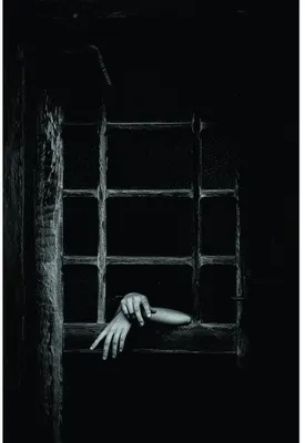 Black and white photograph of two hands reaching through a weathered barred window set into a rough stone wall.