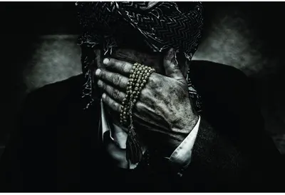 Black and white photograph of aged hands held together in prayer position, wearing rings and holding a beaded strand.