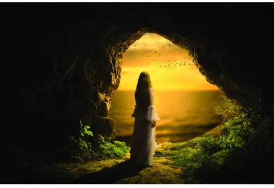A woman in dark clothing stands silhouetted inside a cave, facing a bright golden sunset visible through the cave opening, with green moss covering the rocky interior.