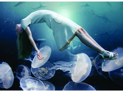 A woman in light-coloured clothing floats upside down underwater, surrounded by glowing jellyfish with translucent bells and trailing tentacles in shades of blue and white.