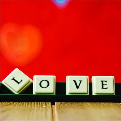 Cream-coloured Scrabble tiles arranged to spell LOVE on a dark wooden surface, with a glowing red heart in the background against a bright red wall.