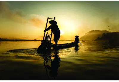 Two silhouetted figures stand in shallow water with boats during a golden sunrise, with mist rising from the water in the background.
