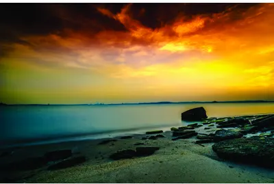 A long-exposure photograph of a rocky beach at sunset, with golden and orange skies above turquoise water and dark rocks in the foreground.