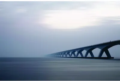 A long concrete bridge with repeating arch supports extends across still blue-grey water under an overcast sky.