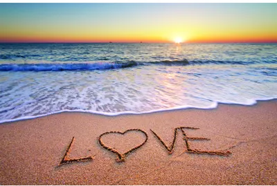 The word 'LOVE' is written in the sand of a beach with a heart symbol replacing the letter O, with ocean waves and a sunset sky in the background.