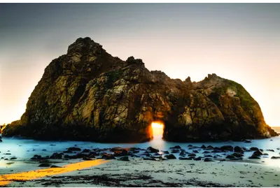 A large rocky island with a natural stone arch through its centre, with golden sunlight streaming through the opening onto dark blue water and rocky shore below.