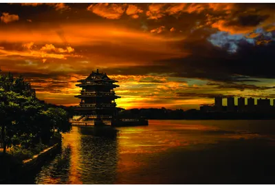 A traditional Asian pagoda silhouetted against an orange and golden sunset sky, reflected in calm water below with dark clouds overhead.