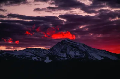 A snow-covered mountain silhouette against a dramatic sunset sky in shades of deep red, purple and gold.