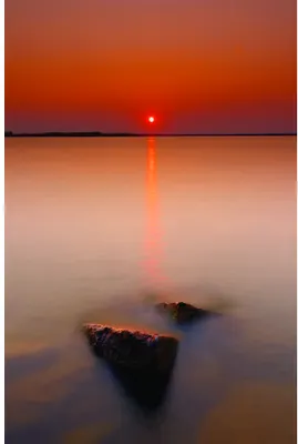 A large sun sets low over still water with a dark rock formation in the foreground, casting a golden reflection across the calm surface.