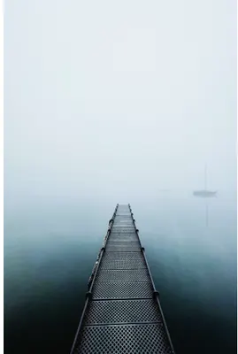 A long metal pier with geometric mesh flooring extends straight into dense fog over calm water, with a sailboat barely visible in the mist.