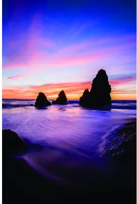 Silhouetted rock formations rise from turbulent purple-blue ocean waters beneath a sunset sky of pink, orange and deep blue.