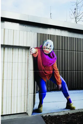 A street art mural of a skateboarder wearing a purple helmet and colourful athletic wear in a low crouch, hand extended, against an urban industrial backdrop.