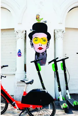 Street art style portrait of a stylised face with yellow eyes, red lips, and a black top hat, with a small yellow bird on top, against a white brick wall background.