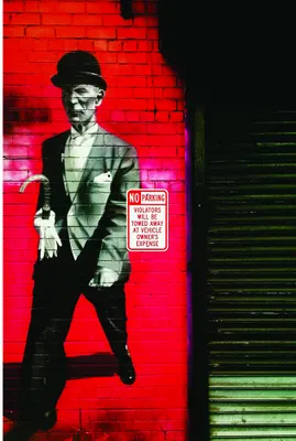 A stylised figure wearing a suit and hat stands against a bright red brick wall illuminated by neon green light.