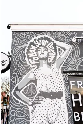Monochrome street art mural of a woman in a polka-dot crop top with her arm raised, set against an intricate psychedelic patterned background.