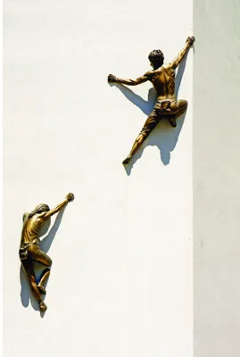 Two bronze-coloured human figures in climbing poses on opposite sides of a white and grey divided wall, casting shadows.