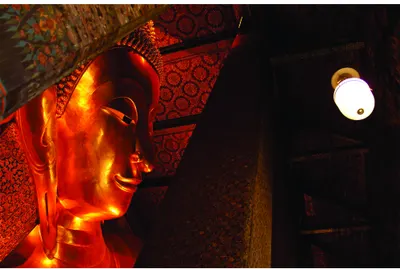 A glowing golden Buddha head is lit from within, surrounded by patterned fabrics and a small white lantern on a dark background.
