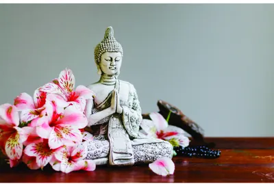 A white Buddha statue sits among bright pink flowers and dark incense sticks on a wooden surface against a grey background.